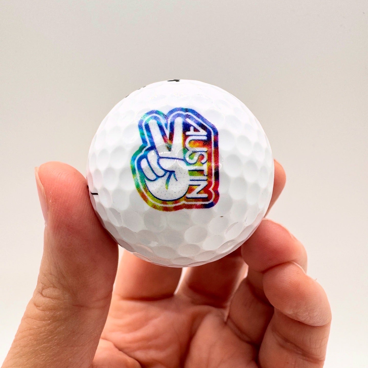 Golf ball with a colorful 'Peace Austin' logo held in a hand against a plain background