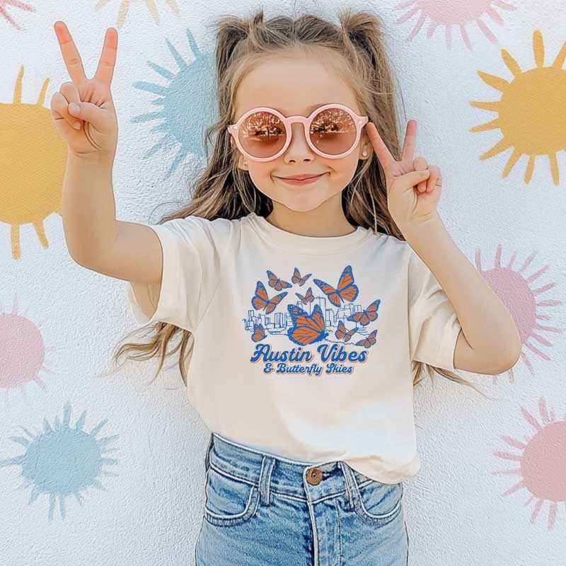 Child wearing a 'Austin Vibes & Butterfly Thins' t-shirt with sunglasses and peace signs, lying on a colorful sun-patterned blanket.