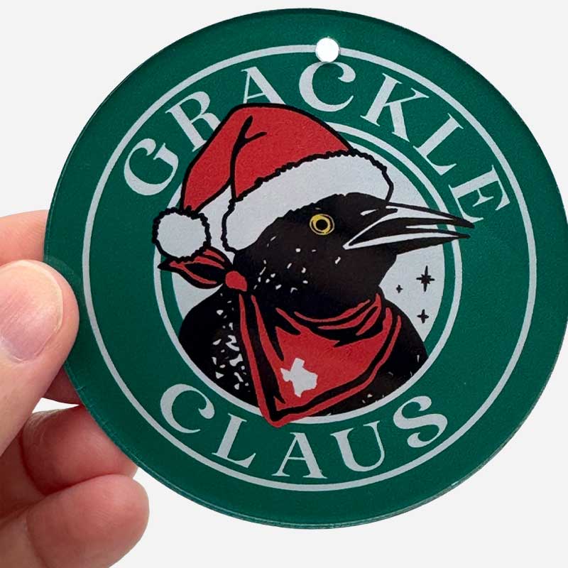 Circular Chiistmas Ornament with a Grackle bird wearing a Santa hat and red scarf, labeled 'Grackle Claus', held by a hand.