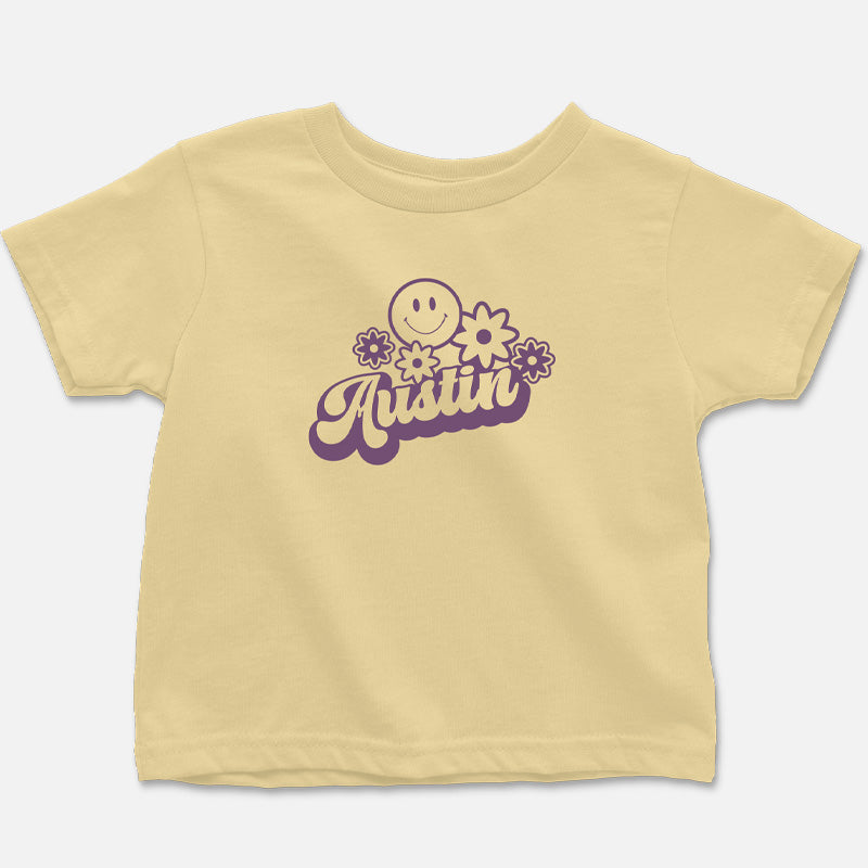 Flat lay of the butter-colored Hippie Austin Toddler Tee with vibrant purple retro lettering, smiley face, and flower design.