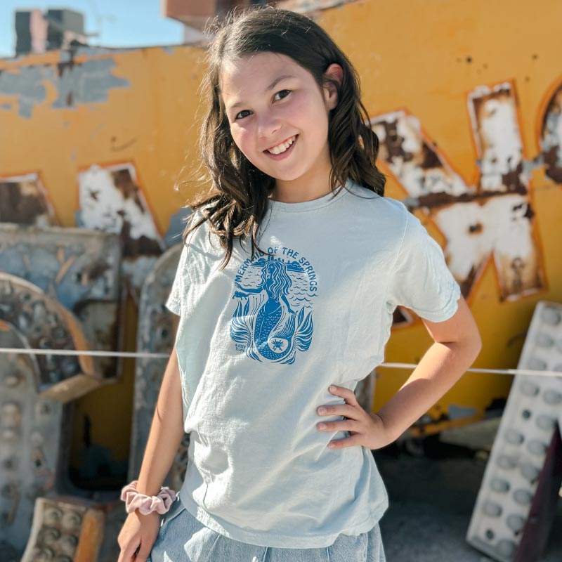 Young girl wearing a light blue t-shirt with a graphic design, standing in front of a colorful mural.