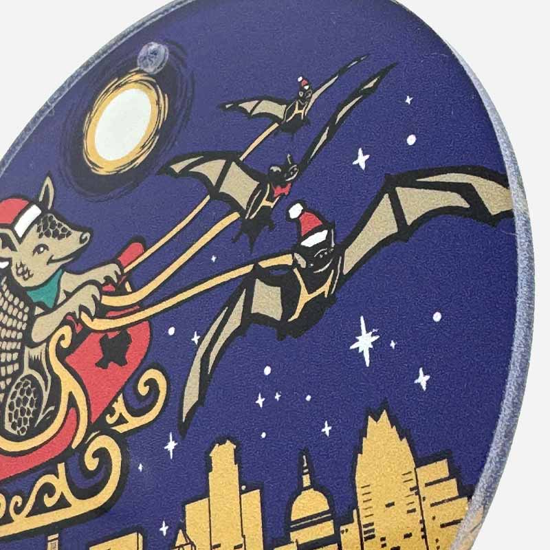 Illustration of a bat in a Santa hat riding in Santas sleight over the Austin, Texas cityscape with a moon and stars in the background.