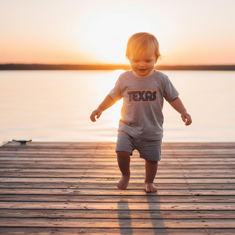 Child wearing a 'Texas' shirt walking on a wooden dock at sunset.