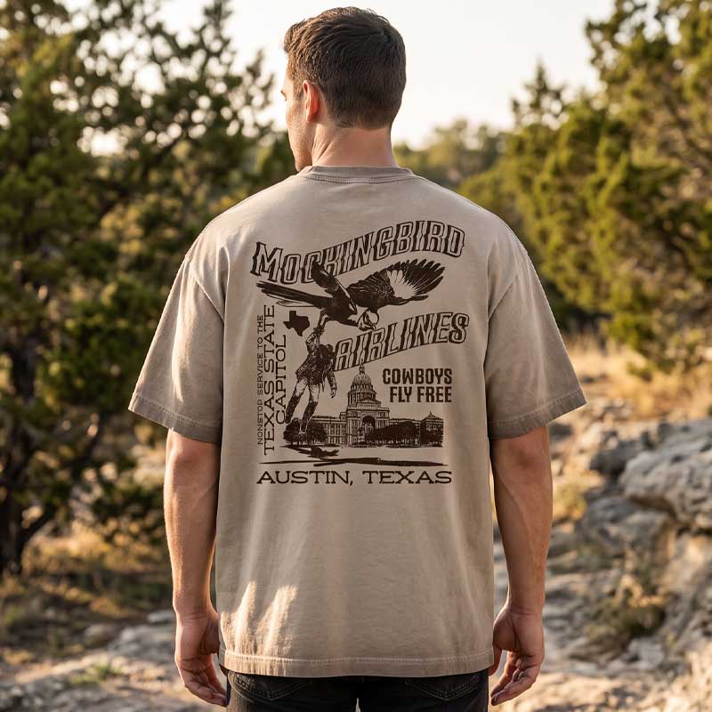 A man viewed from behind wearing a beige graphic t-shirt with a large black print of a mockingbird, an eagle, a cowboy, and the text 'MOCKINGBIRD AIRLINES' and 'AUSTIN, TEXAS' on a natural outdoor background.