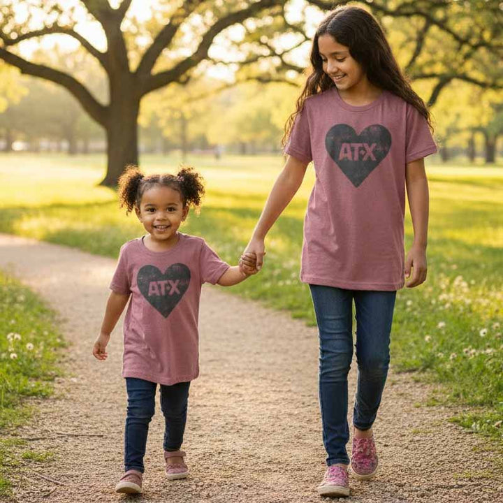 Two young girls walking hand-in-hand on a path in a park, wearing matching pink t-shirts with 'ATX' heart designs.