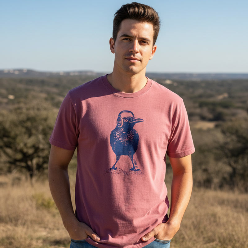 Man wearing a brick comfort colors t-shirt with a blue graphic of a grackle wearing headphones, standing in a scenic outdoor setting.