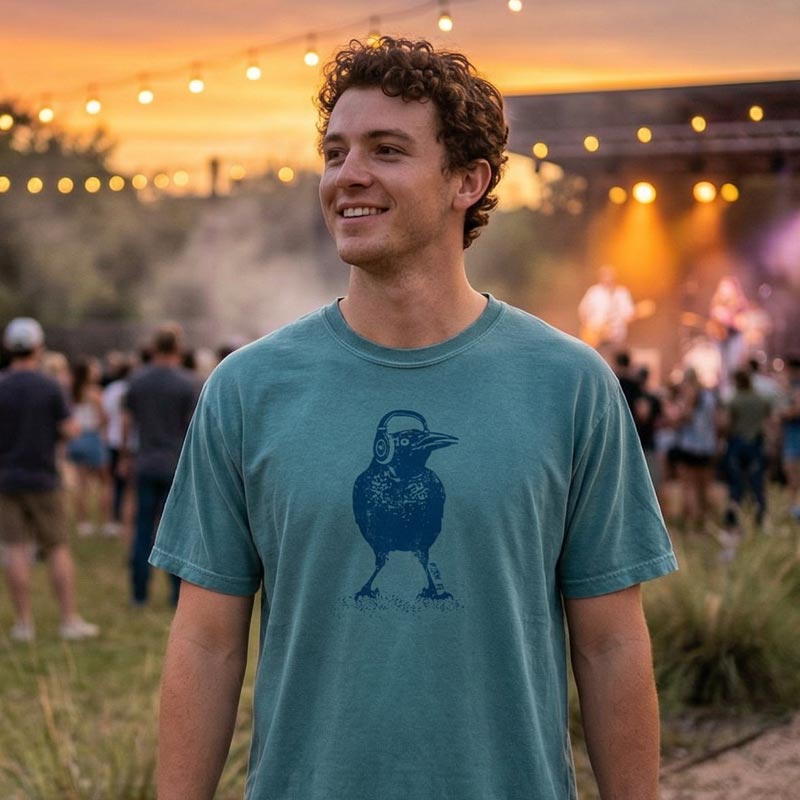 Man wearing a teal t-shirt with a graphic design, standing outdoors with a crowd and lights in the background.