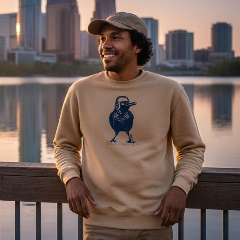 Man wearing a beige sweatshirt with a blue graphic, standing by a waterfront with a city skyline in the background.