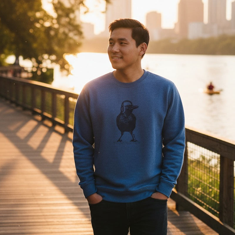 Man wearing a blue sweatshirt with a bird design by a waterfront at sunset.