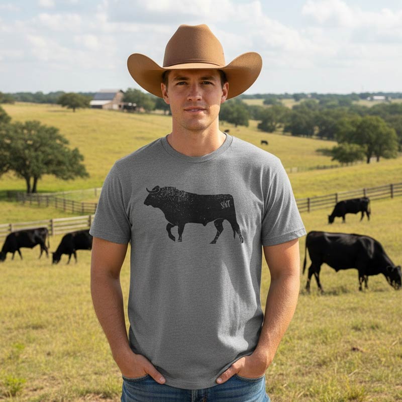 Man wearing a gray t-shirt with a bull graphic and a cowboy hat, standing in a field with cows.