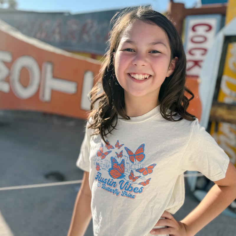 Young girl wearing a t-shirt with butterfly design and text, standing in front of vintage signs.