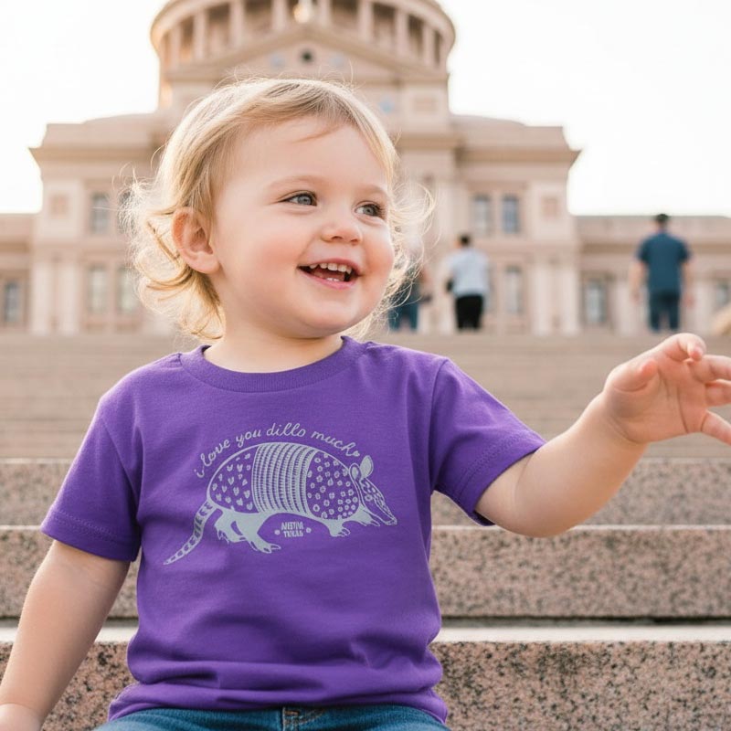 Child wearing a purple t-shirt with an armadillo graphic in front of a building.