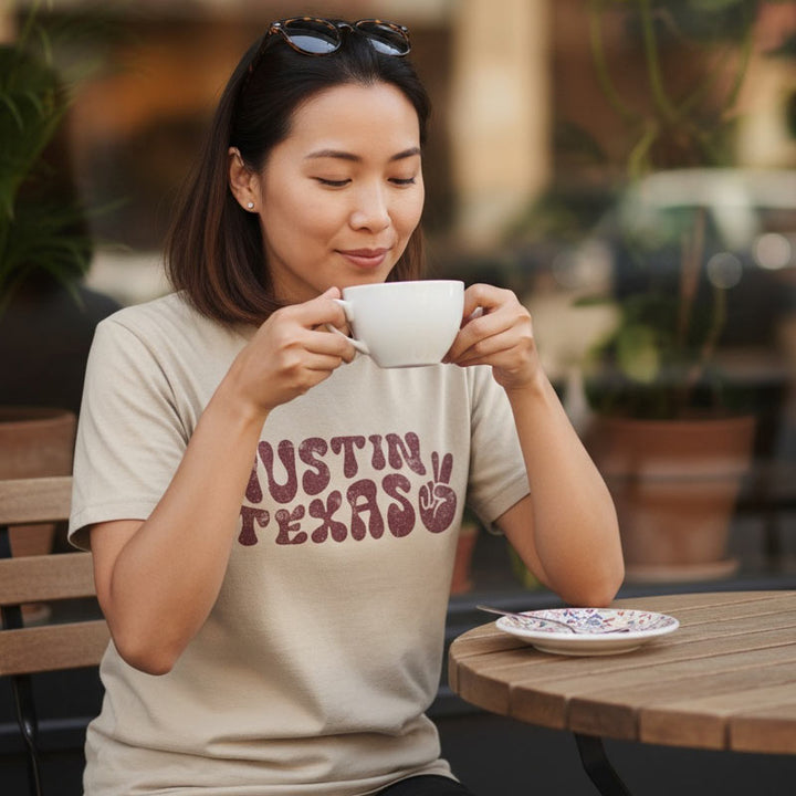 Woman drinking from a cup at an outdoor cafe wearing a 'Austin, Texas' t-shirt.