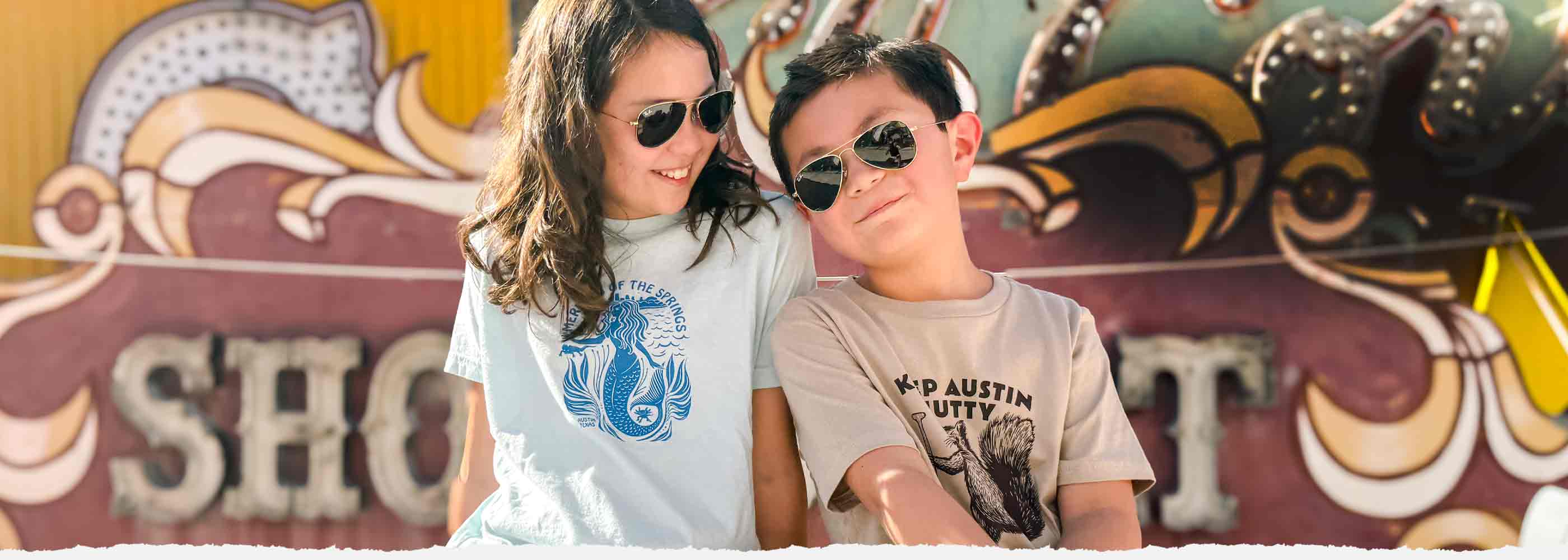 Two kids wearing sunglasses in front of a colorful backdrop wearing Gusto Graphic Tees Youth designs