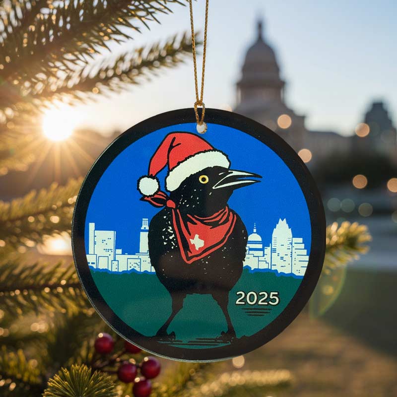 Christmas ornament with a Grackle bird wearing a Santa hat and scarf, set against the Austin, Texas skyline with '2025' text, on a Christmas tree.