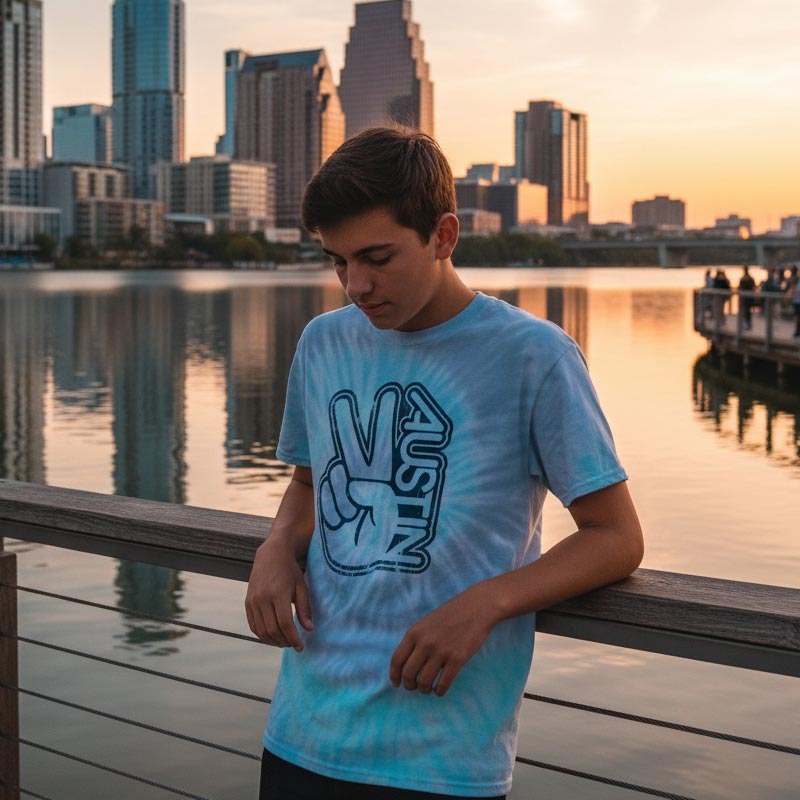 Person wearing a blue tie-dye t-shirt with a peace sign design by a waterfront at sunset.