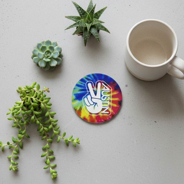 Colorful tie-dye coaster with peace symbol and 'Austin' text on a gray surface with plants and a mug.