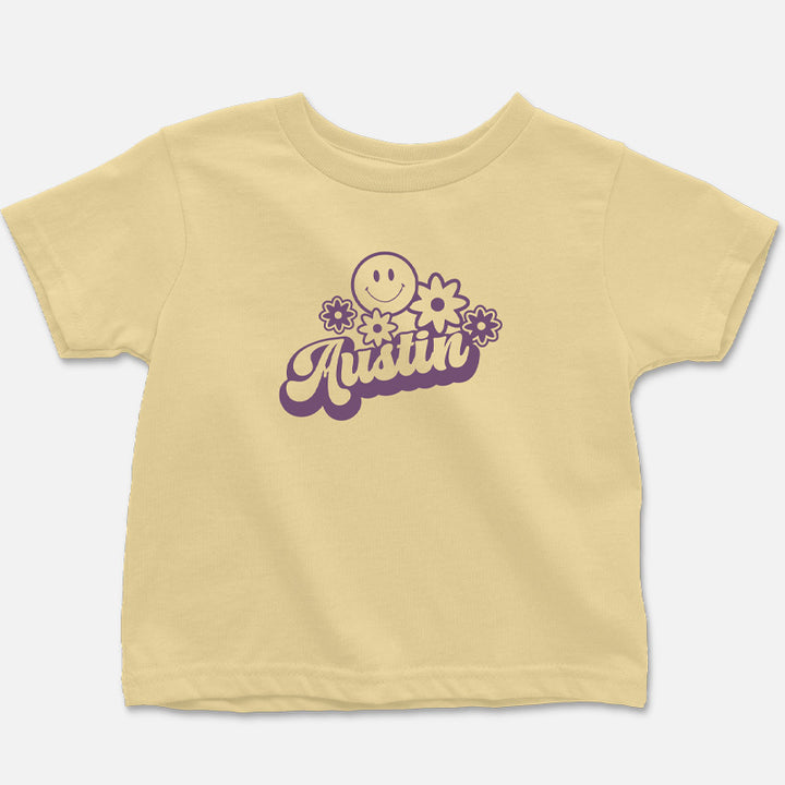 Flat lay of the butter-colored Hippie Austin Toddler Tee with vibrant purple retro lettering, smiley face, and flower design.