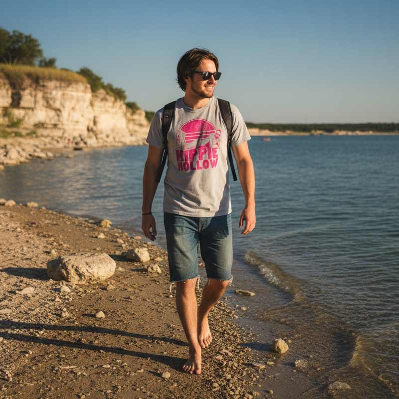 Man walking along a rocky shoreline with a backpack and 'Hippie Hollow' t-shirt.