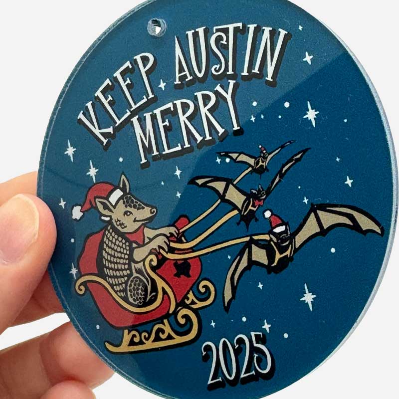 Circular ornament with 'Keep Austin Merry' text, cartoon reindeer, and bat, held by a hand.