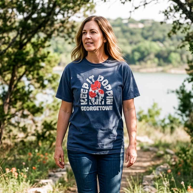 Woman wearing a navy Keep it Poppin, Georgetown, Texas  t-shirt, standing outdoors near a body of water.