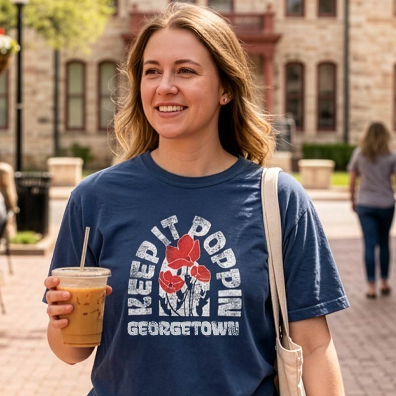 Woman wearing a navy Keep It Poppin Georgetown, Texas t-shirt with a graphic design, holding a drink, and standing on a sidewalk.