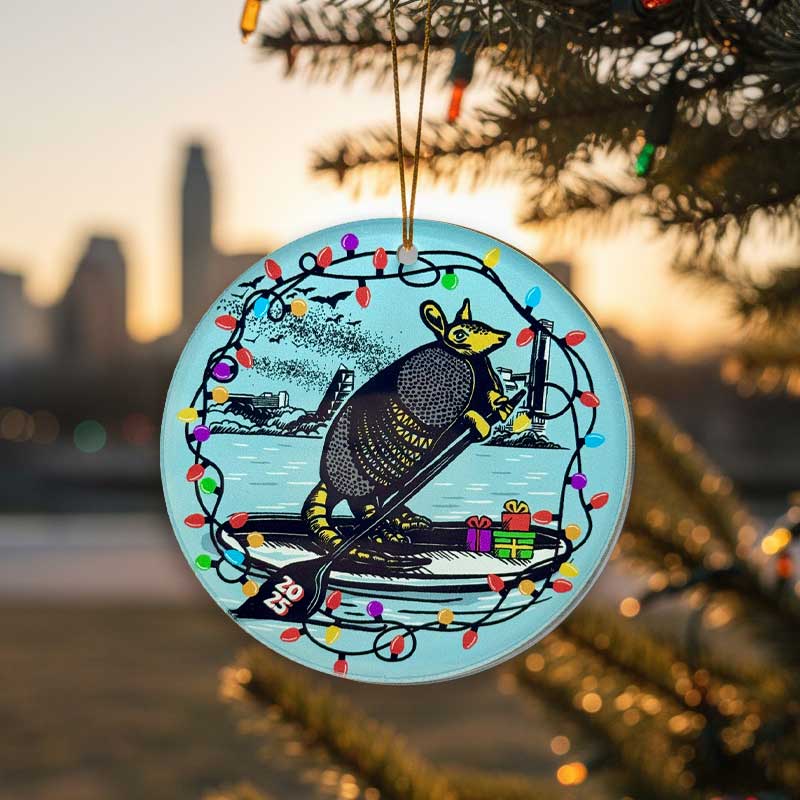 Decorative Christmas ornament with Festive armadillo paddle boarding on Lady Bird Lake with a Christmas light border.