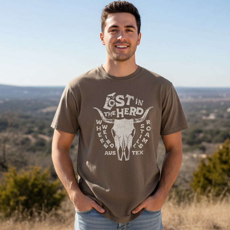 Man wearing a Lost In The Herd Austin, Texas brown t-shirt with a graphic design standing outdoors with a scenic background
