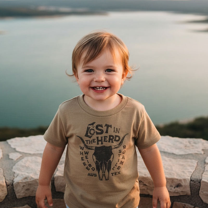 Child wearing a 'Lost in the Hero' t-shirt with a skull design, standing by a scenic lake.