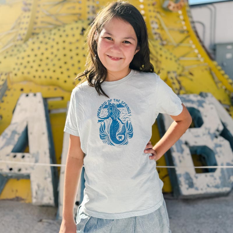 Young girl wearing a t-shirt with a blue mermaid design in front of a large, colorful sign.