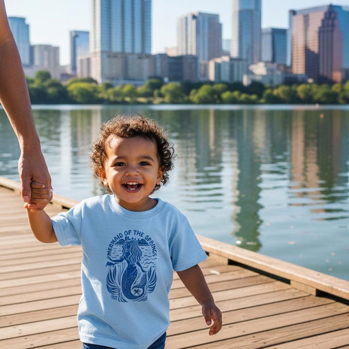 Child holding hands with an adult on a wooden dock by a lake with city skyline in the background