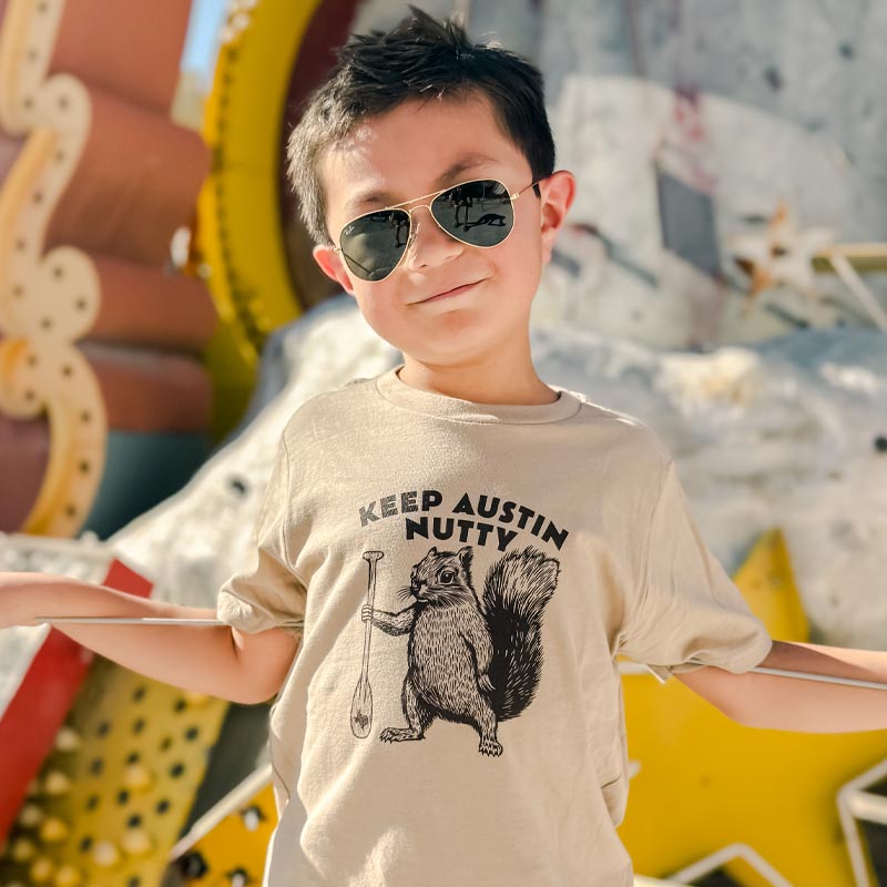 Child wearing sunglasses and a Keep Austin Nutty t-shirt with a graphic of a Squirrel in an outdoor setting