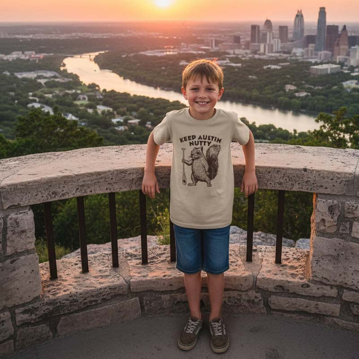 Child wearing a 'Keep Austin Nutty' t-shirt standing on a stone ledge with a cityscape and river in the background.