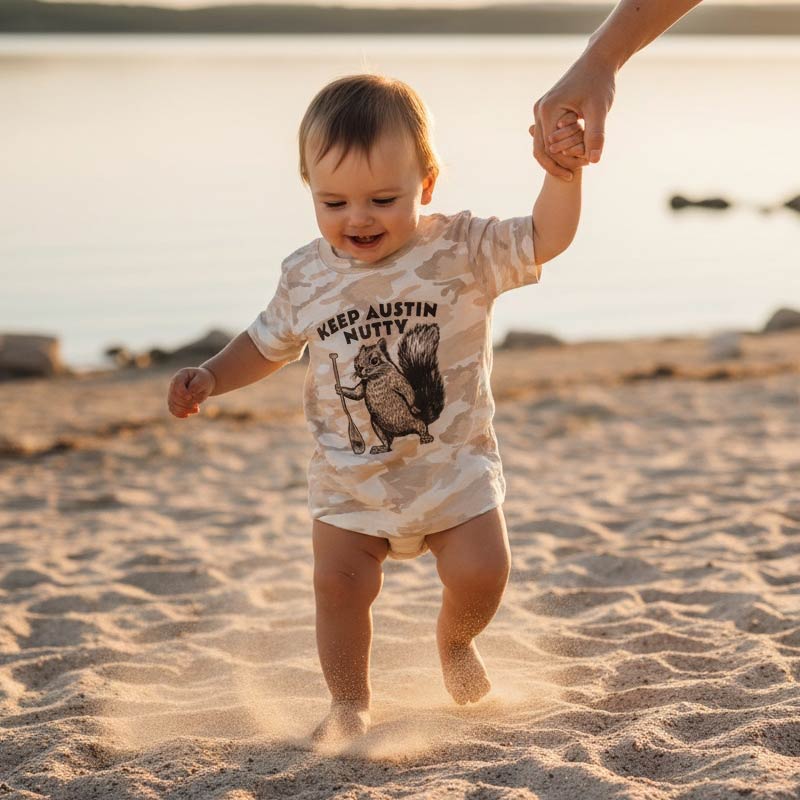 Child in a 'Keep Austin Nutty' shirt walking on a sandy beach with an adult's hand.