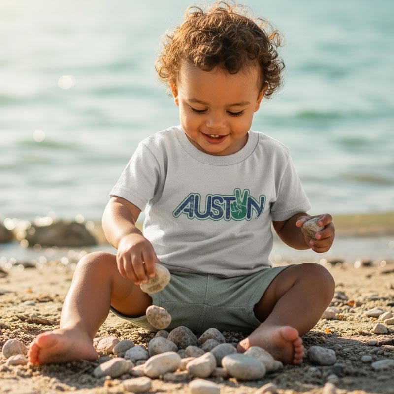 Child playing with pebbles on a beach wearing an 'AUSTON' shirt.