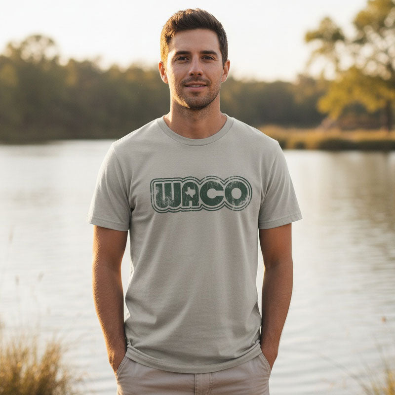 Man wearing a 'Waco' t-shirt standing by a lake with trees in the background
