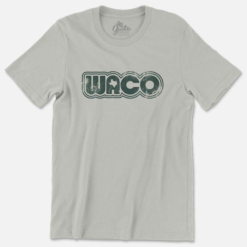 Unisex soft thyme green Retro Waco graphic T-shirt. Design features bold retro lines spelling “Waco” with a tiny Texas silhouette inside the “A.”