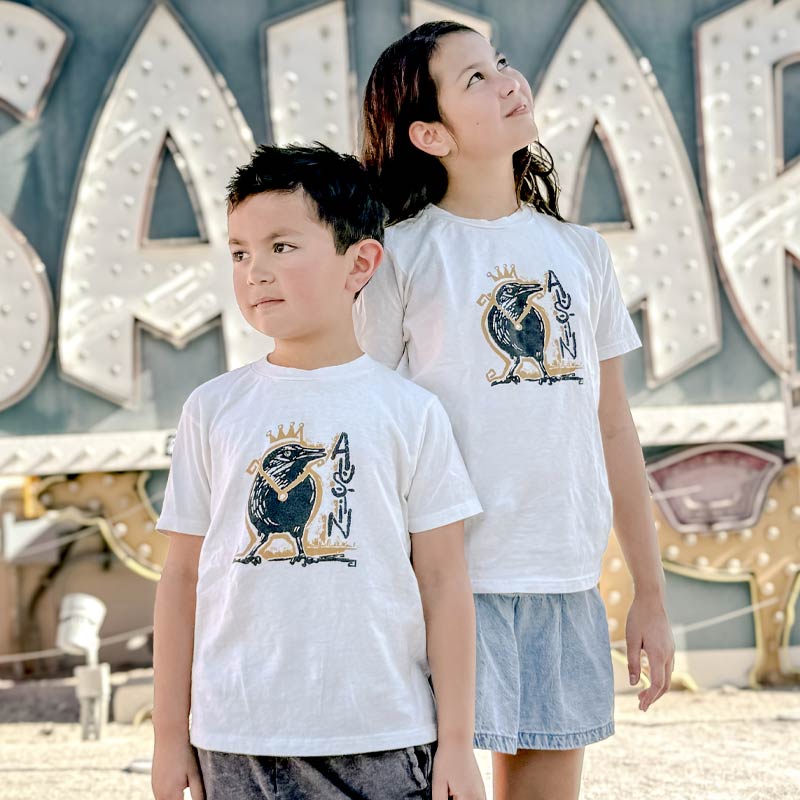 Two children wearing white t-shirts with a logo in front of a neon sign.