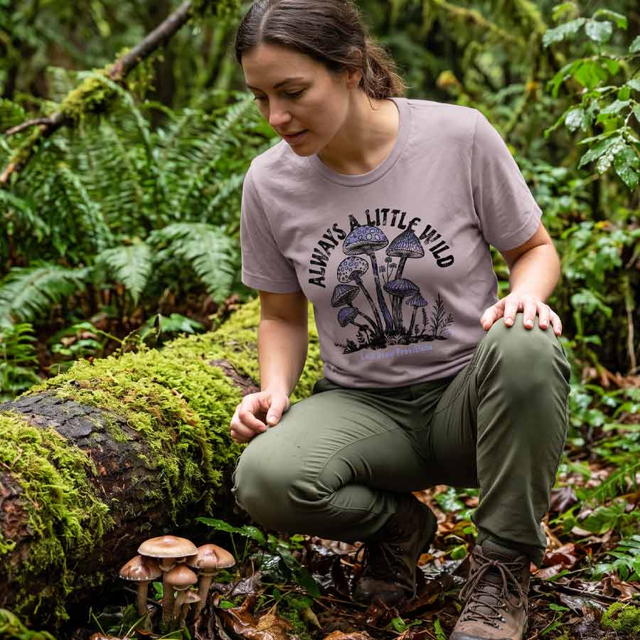A woman crouching in a lush green forest next to a mossy log and small mushrooms, wearing a taupe graphic t-shirt that reads 'ALWAYS A LITTLE WILD' with a mushroom design and olive green pants.