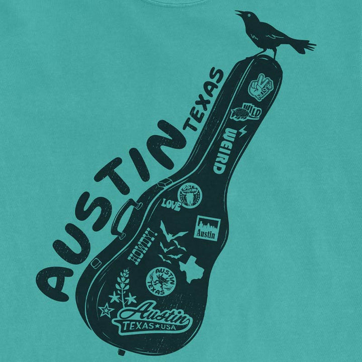 Close up view of a Seafoam Comfort Colors 1717 Carry-On Austin t-shirt laid flat on a neutral background. Features a black front-only graphic of a sticker-covered guitar case with Austin-inspired artwork and a grackle perched on top.