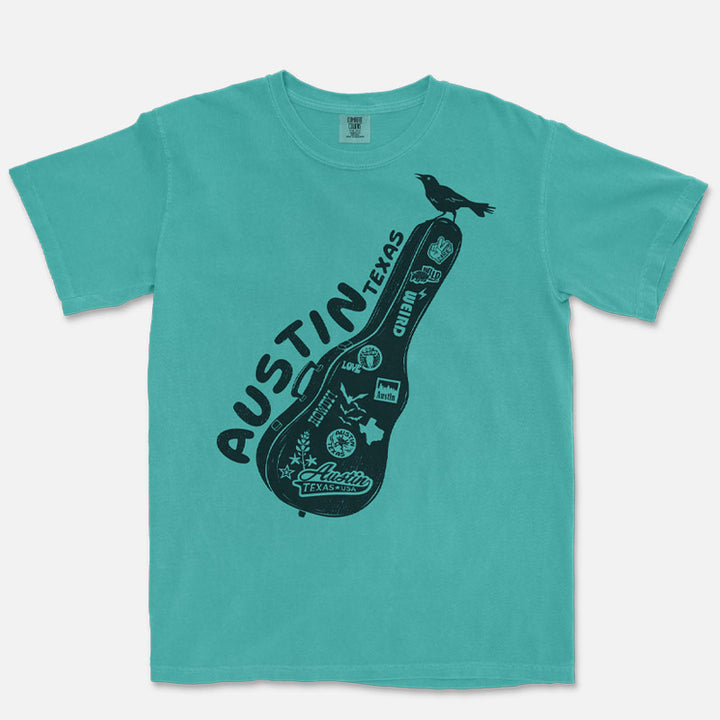 Front view of a Seafoam Comfort Colors 1717 Carry-On Austin t-shirt laid flat on a neutral background. Features a black front-only graphic of a sticker-covered guitar case with Austin-inspired artwork and a grackle perched on top.