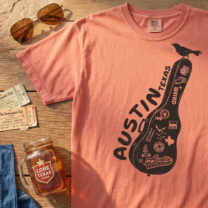 Terracotta Comfort Colors 1717 Carry-On Austin t-shirt laid flat with Austin-inspired accessories including sunglasses, guitar picks, and travel-inspired details. Front graphic features a guitar case covered in Austin stickers and a grackle.