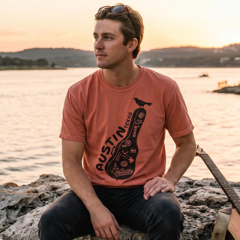 Man wearing a terracotta Carry-On Austin t-shirt outdoors near the water, styled with a casual Texas look. Front-only Austin guitar case graphic clearly visible.