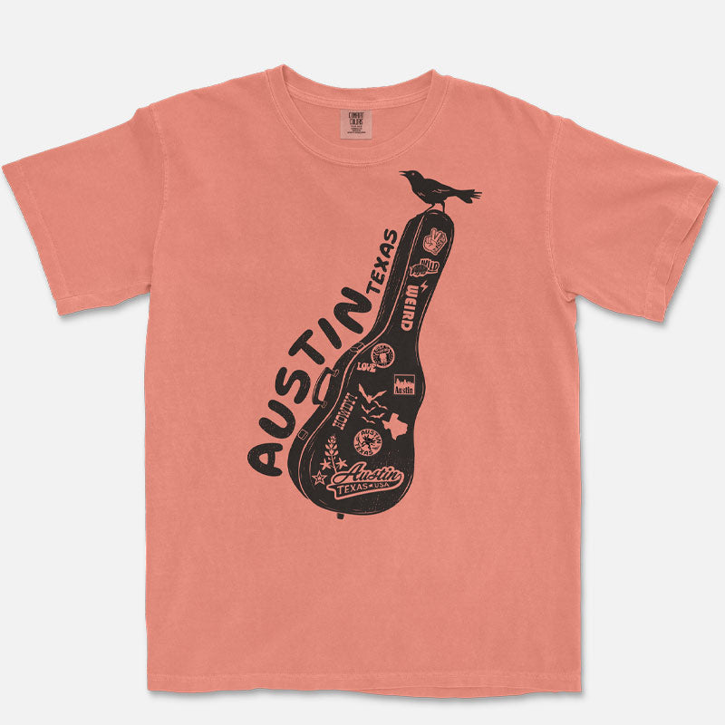 Front view of a terracotta Comfort Colors 1717 Carry-On Austin t-shirt laid flat on a neutral background. Features a black front-only graphic of a sticker-covered guitar case with Austin-inspired artwork and a grackle perched on top.