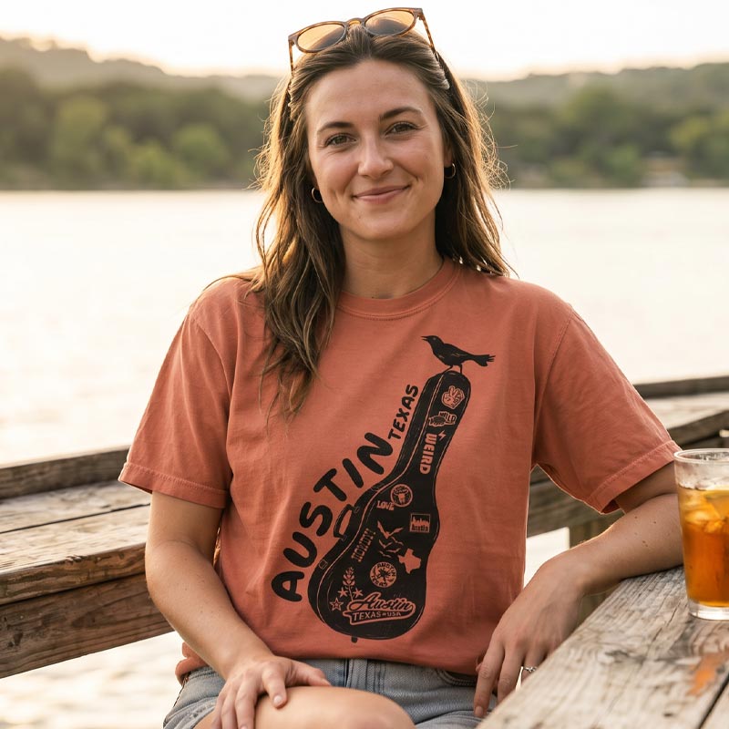 Woman relaxing lakeside in a terracotta Carry-On Austin graphic tee, featuring a front-only guitar case design with Austin-inspired artwork and a grackle. Warm golden light and relaxed Austin summer vibe.