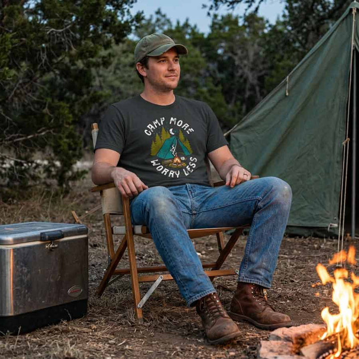 A man sits in a folding camp chair outdoors near a tent and campfire, wearing a dark graphic T-shirt with the text &