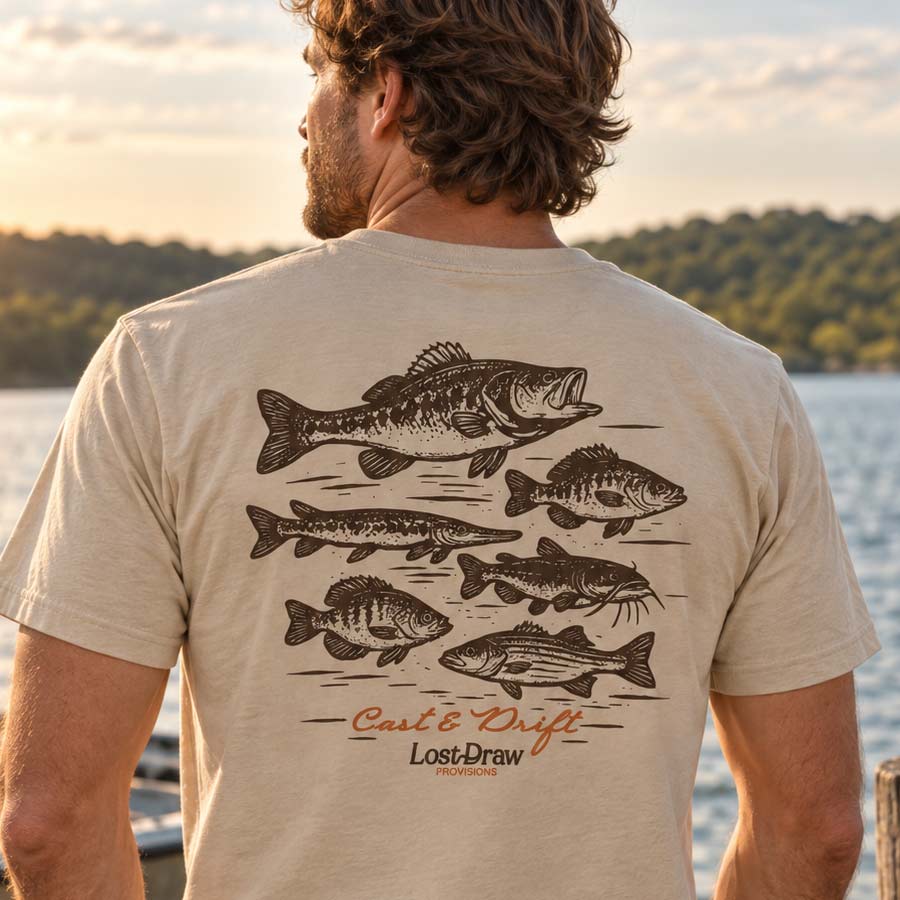 A man viewed from behind wearing a beige t-shirt with a large black fish graphic and 'Cast & Drift Lost Draw Productions' printed on the back, set against a scenic lake background at sunset.