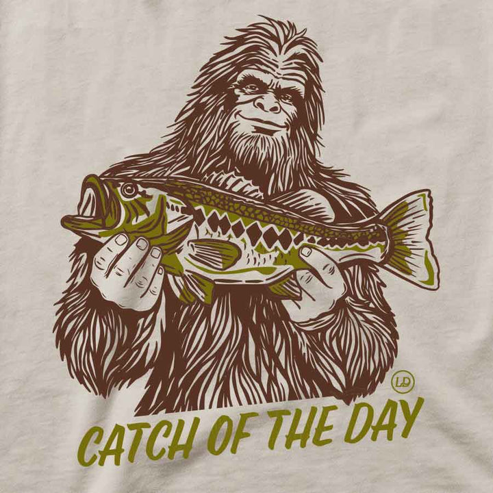 A close-up of a graphic t-shirt featuring a brown line art illustration of Bigfoot holding a green and brown fish, with the text &
