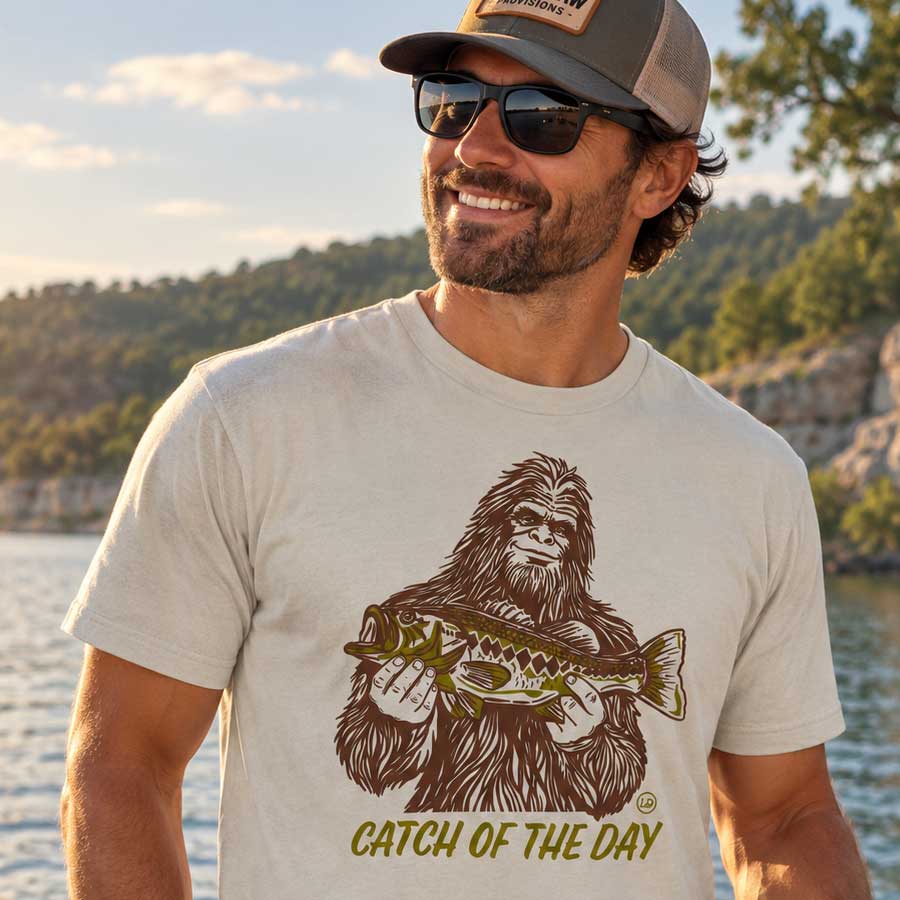 A man wearing a beige graphic t-shirt with a brown and olive-green print of Bigfoot holding a fish and the text &