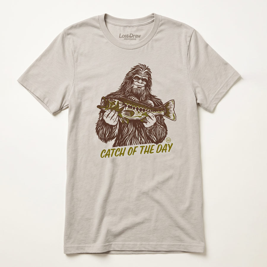 A light beige crewneck t-shirt featuring a graphic of Bigfoot holding a fish, with the text &